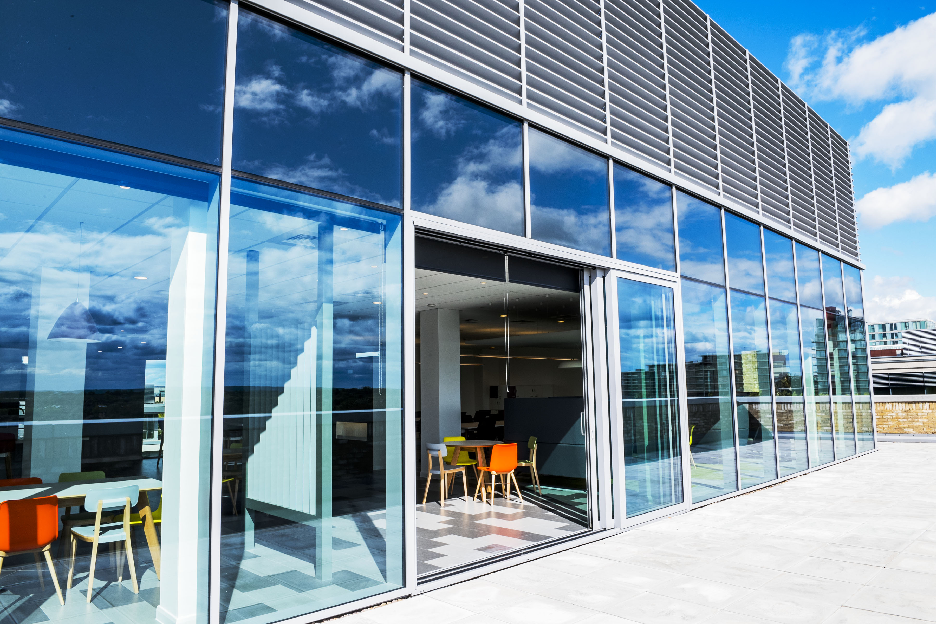 Photos shows the view from the balcony into the Milton Keynes office. A glass building, with the doors open, and a bright airy office space inside. 