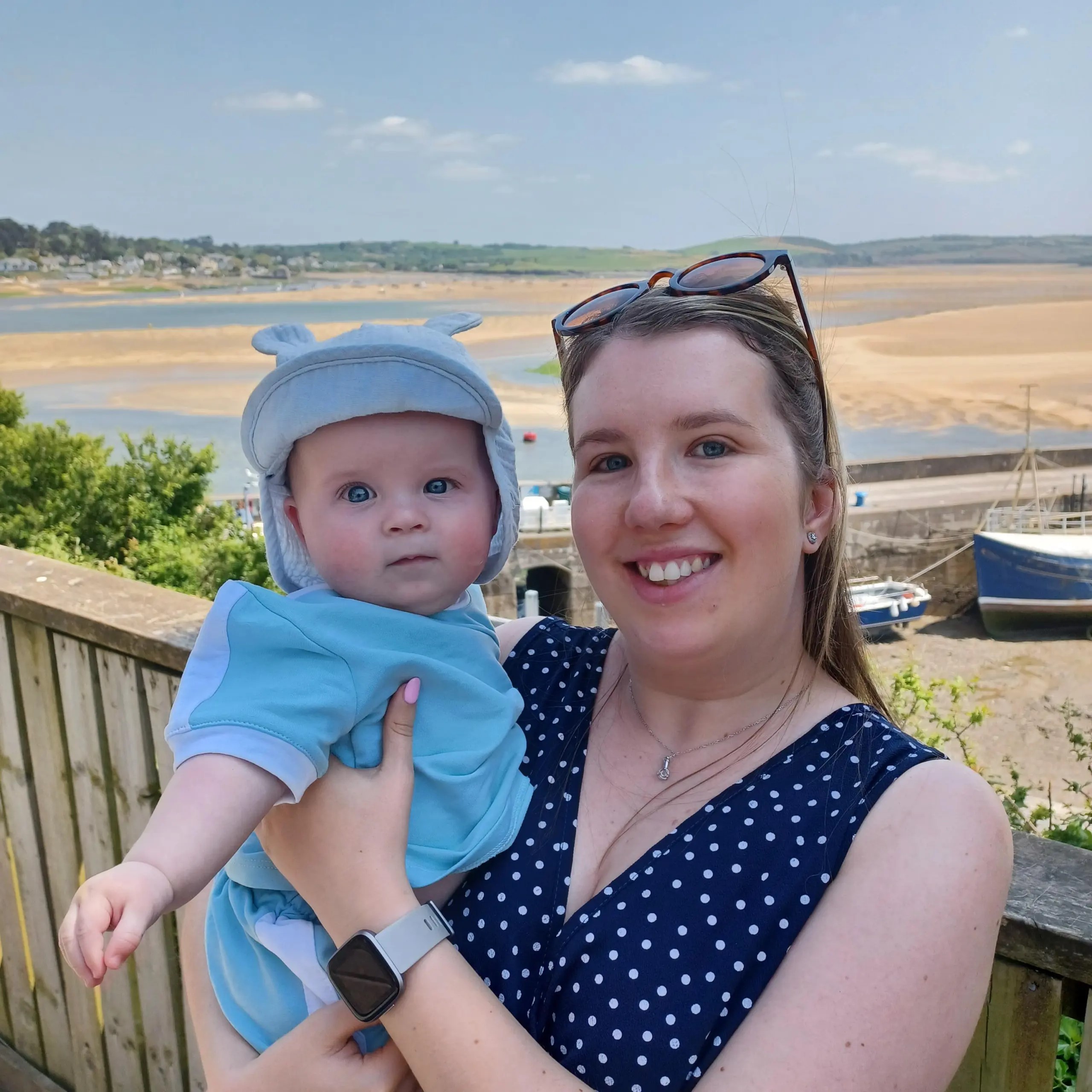 Emma is wearing a blue and white summer dress, holding her child in her arms. They have the beach in the background and are smiling at the camera.